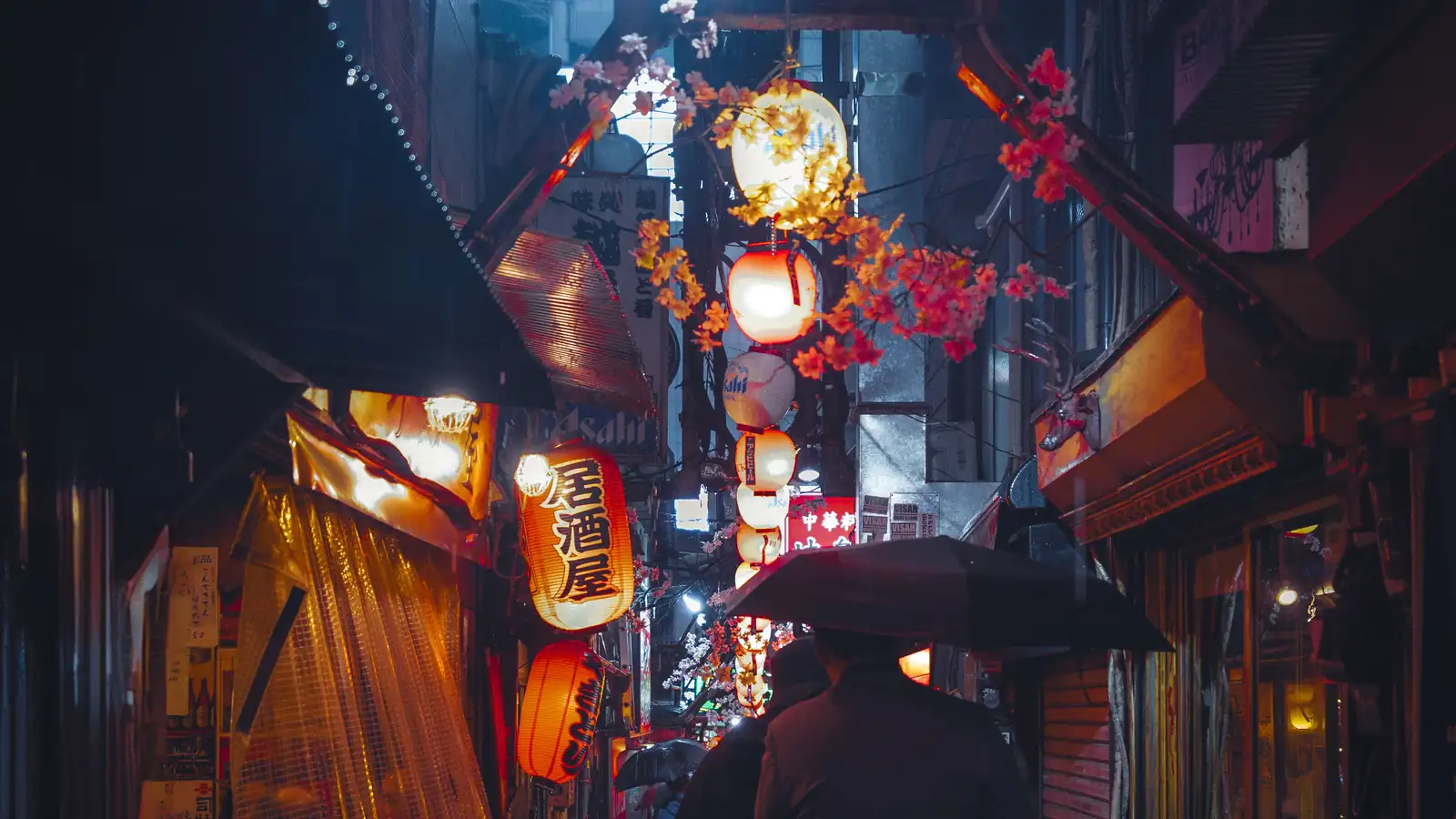 A narrow alley at night in the rain, lit by red and white paper lanterns, with a person under an umbrella walking away