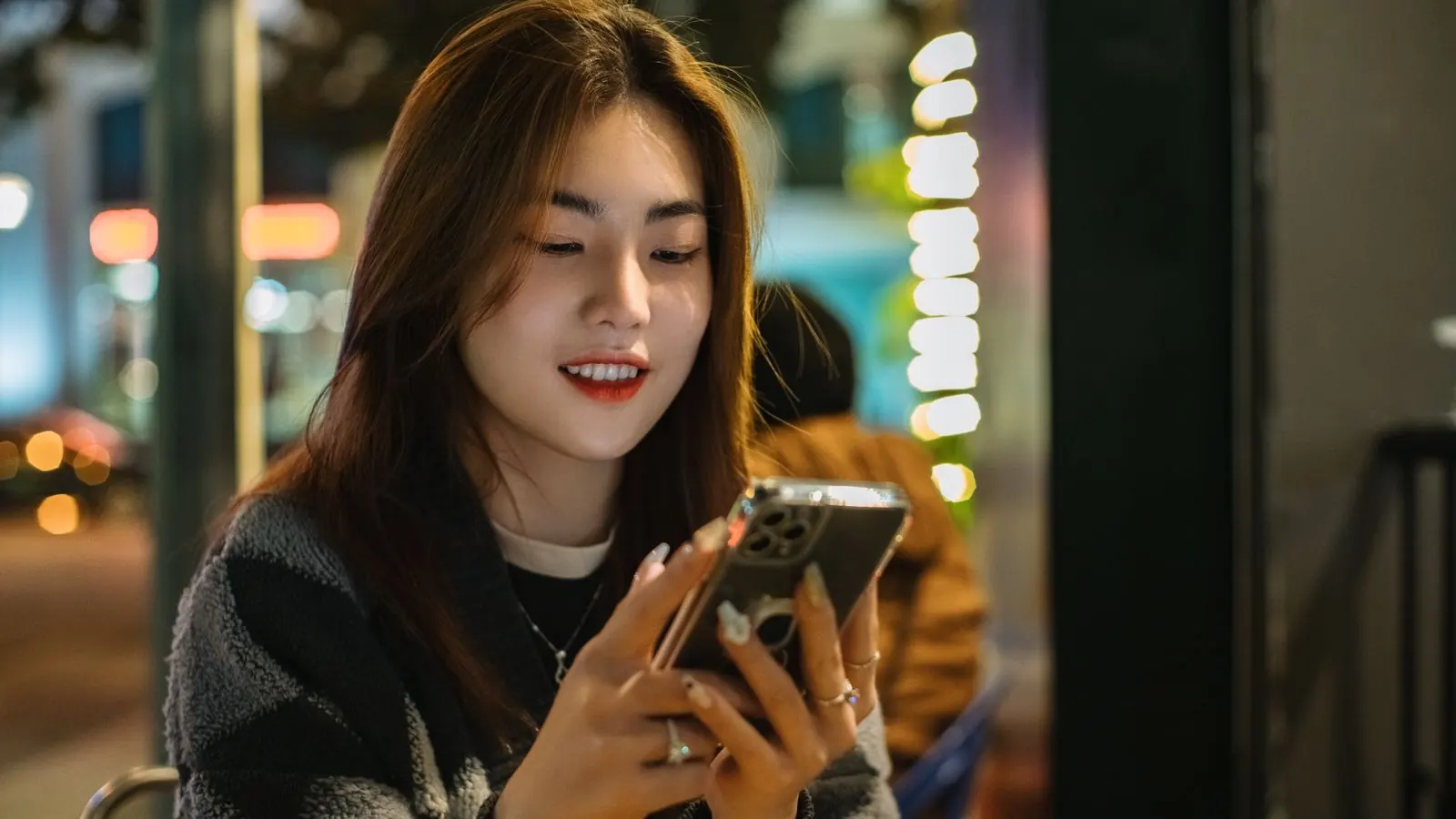 A smiling woman sitting outdoors in the evening, texting on her phone