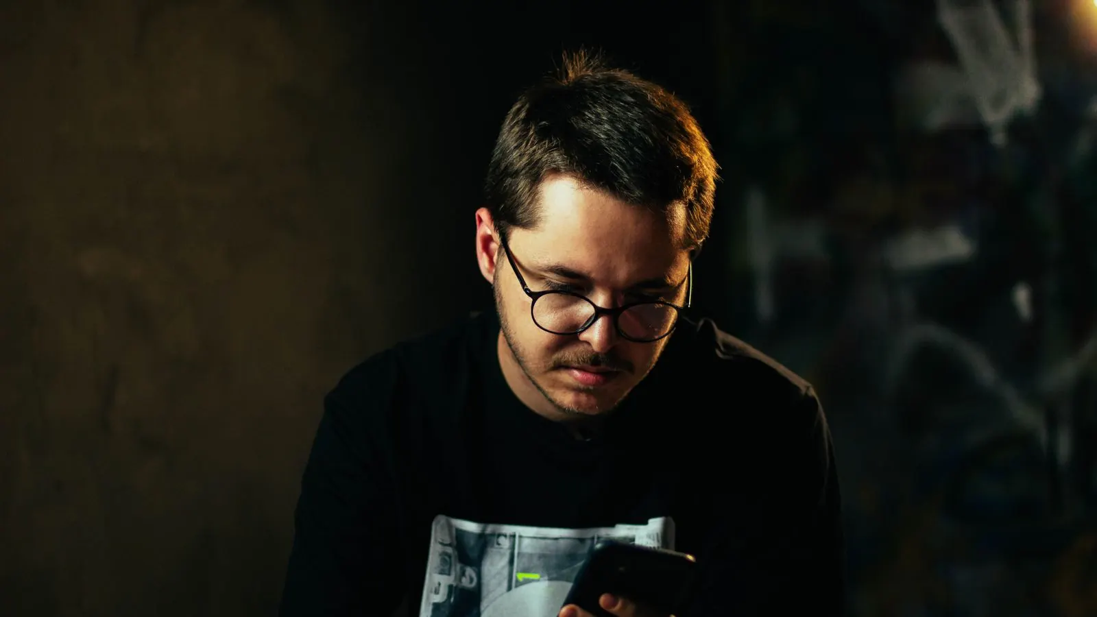 A man in glasses reading his smartphone in a dim, moody room