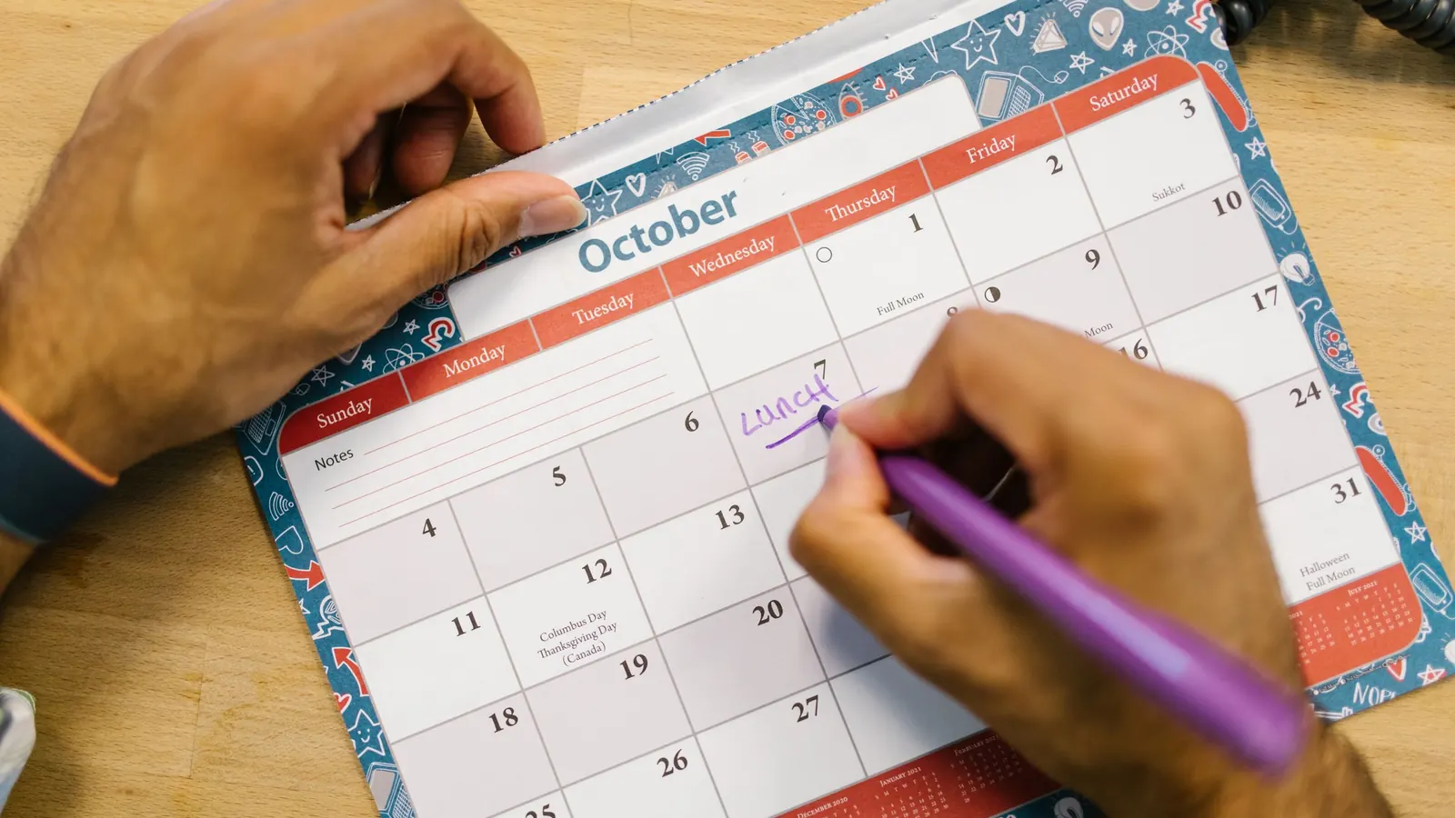 Close-up of a hand writing a reminder on an October desk calendar with a purple pen