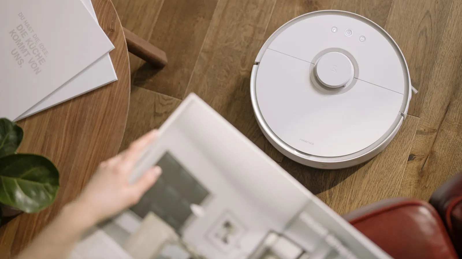 A white round robot vacuum with a laser turret crossing a dark wooden floor beside a side table and an open magazine