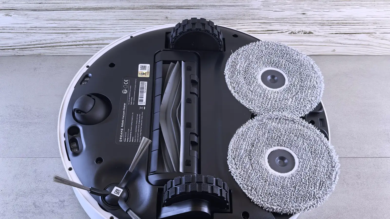 The underside of a robot vacuum and mop showing two round mop pads, a roller brush, a side brush and two rubber wheels